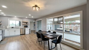 A spacious kitchen with white cabinets, a dining area with a long table and chairs, and large windows offering a view of the backyard. - Excellent location, close to Olympic National Park, restaurants & grocery stores (Port Angeles)