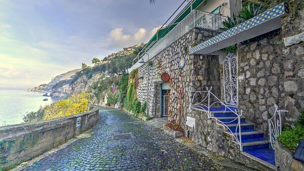A coastal street with stone buildings, a cobblestone path, and a staircase leading to a balcony overlooking the sea.