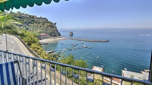 A coastal view from a balcony overlooking a marina with boats, a rocky coastline, and buildings on a hill.