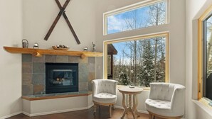 A fireplace with stone tiles, two upholstered chairs, a small round table, and a large window with a view of trees.