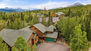 A cluster of log cabin-style buildings surrounded by dense forest, with a mountain range in the background. - 113 Burgundy Circle by Summit County Mountain Retreats (Silverthorne)