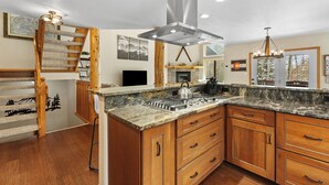 A kitchen with a granite countertop, wooden cabinets, a stainless steel range hood, and a wooden staircase.