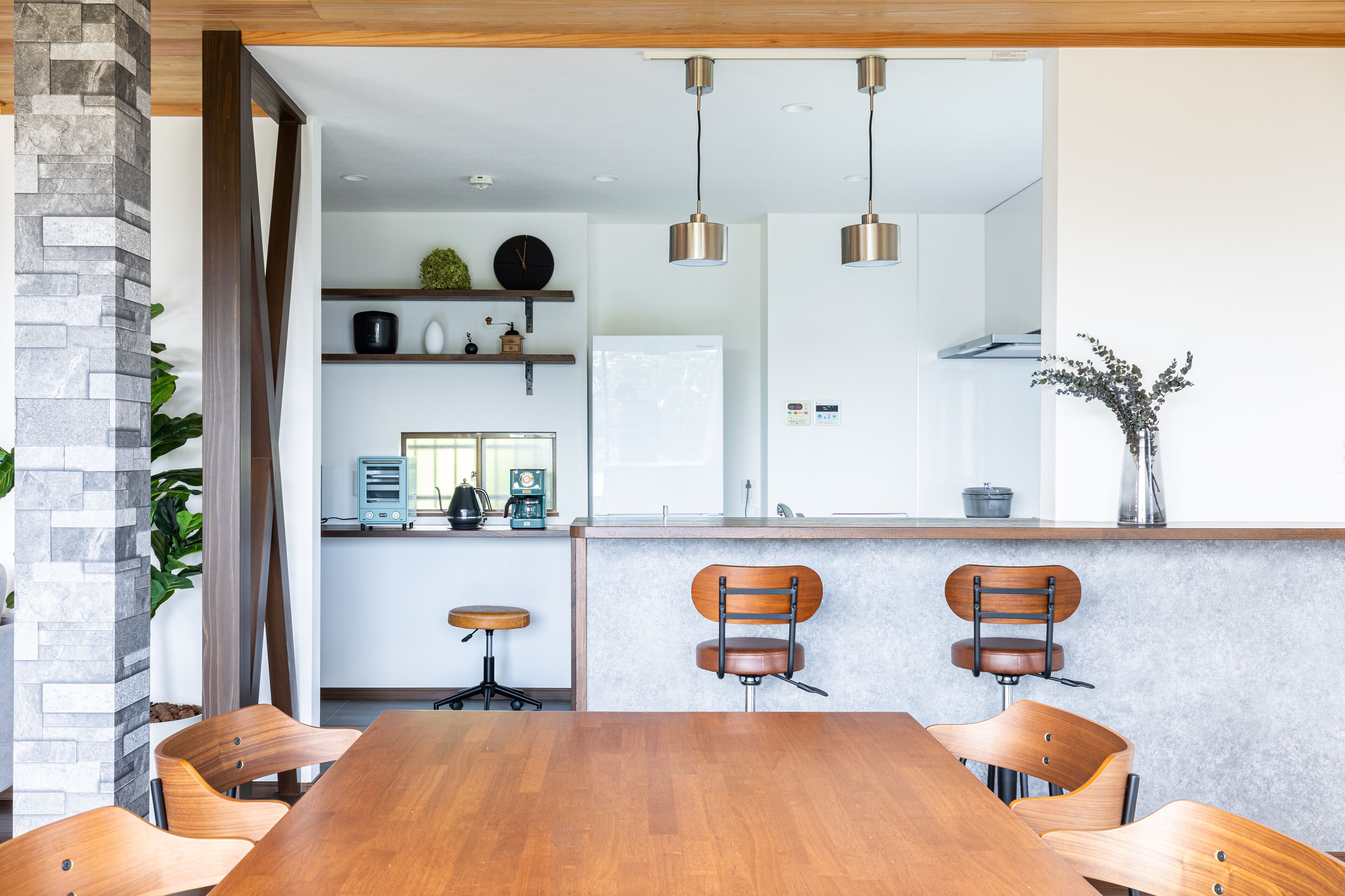 A modern kitchen with a central island, wooden chairs, pendant lights, and a stone pillar.