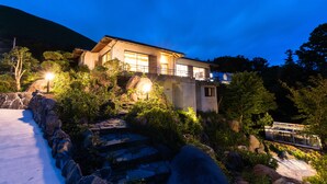 A modern house with a stone pathway leading to the entrance, surrounded by large rocks and greenery. - Resort stay Bowen (Ito)
