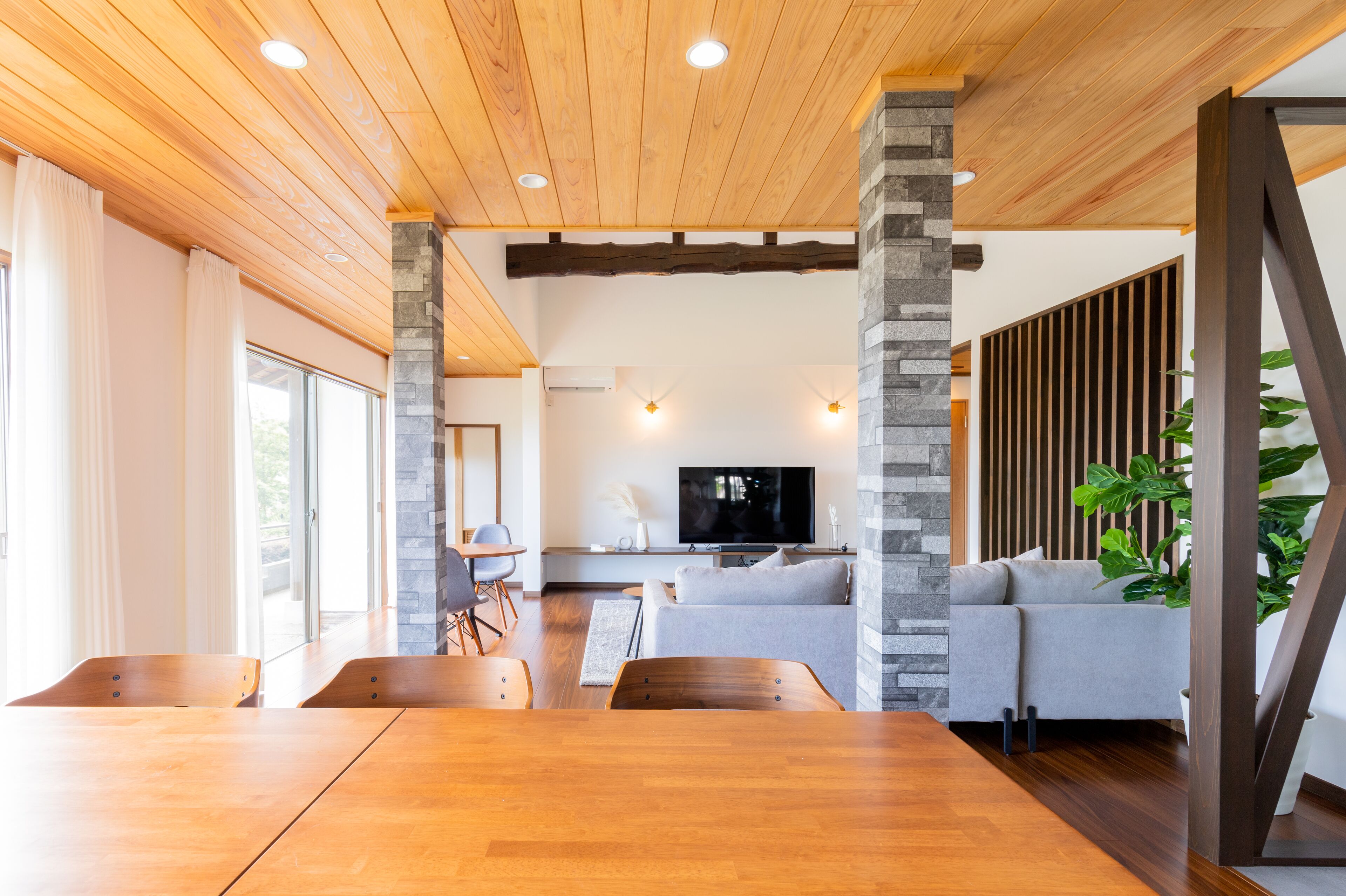Una sala de estar moderna con techo de madera, pilares de piedra, un sofá gris, un televisor y un comedor con mesas y sillas de madera.