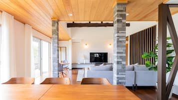 Una sala de estar moderna con techo de madera, pilares de piedra, un sofá gris, un televisor y un comedor con mesas y sillas de madera.