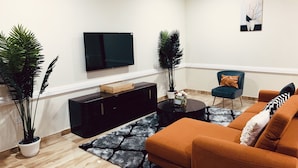 A modern living room with a flat-screen TV, a black entertainment unit, a round coffee table, an orange sofa, and two potted plants. - Modern and Spacious 2 Bdrm Serviced Apt w/ Patio - Approx. 15 mins from Airport (Accra)
