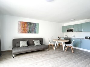 Een moderne keuken met blauwgroene kastjes, een zithoek met een grijze bank en gestreepte kussens, en een kleurrijk abstract schilderij aan de muur.