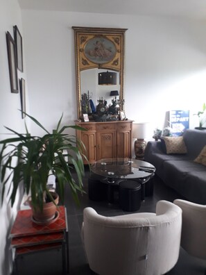 A living room with a round glass table, a sofa, a chair, a potted plant, a wooden cabinet with a mirror, and a clock.