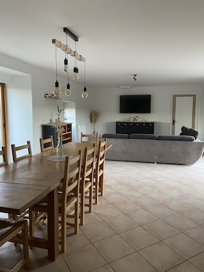 Ein Essbereich mit Holztisch und Stühlen, einem an der Wand befestigten Fernseher und einer Couch im Hintergrund.