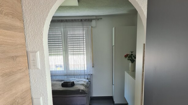 En smal hall med en säng, ett fönster med persienner, en liten växt och en dörr.