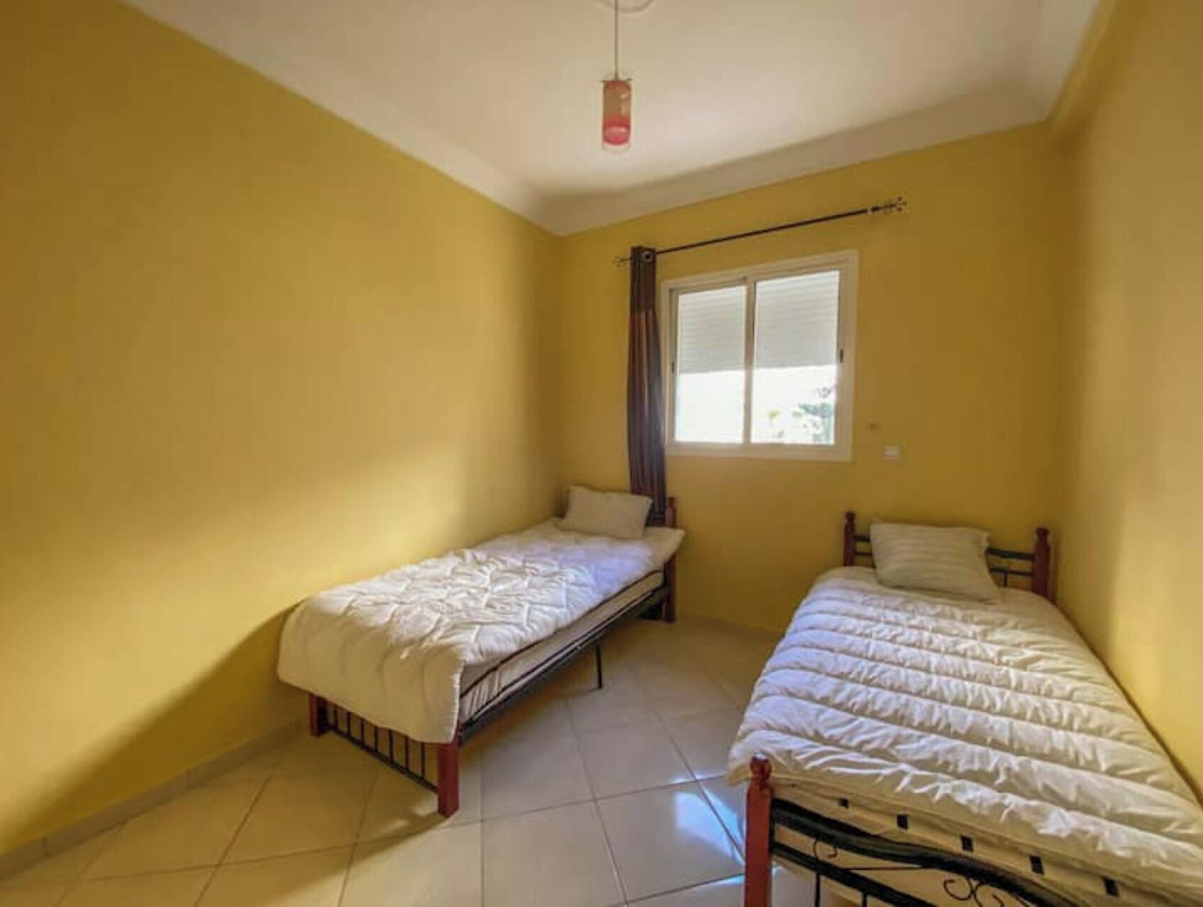 Une chambre avec deux lits simples, une fenêtre avec des stores et un luminaire suspendu.