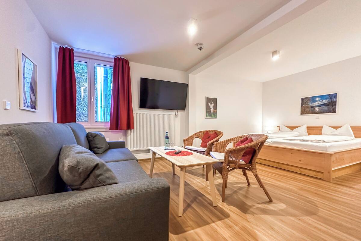 A modern living room with a grey sofa, a wooden coffee table, wicker chairs, a wooden bed, and a TV mounted on the wall.