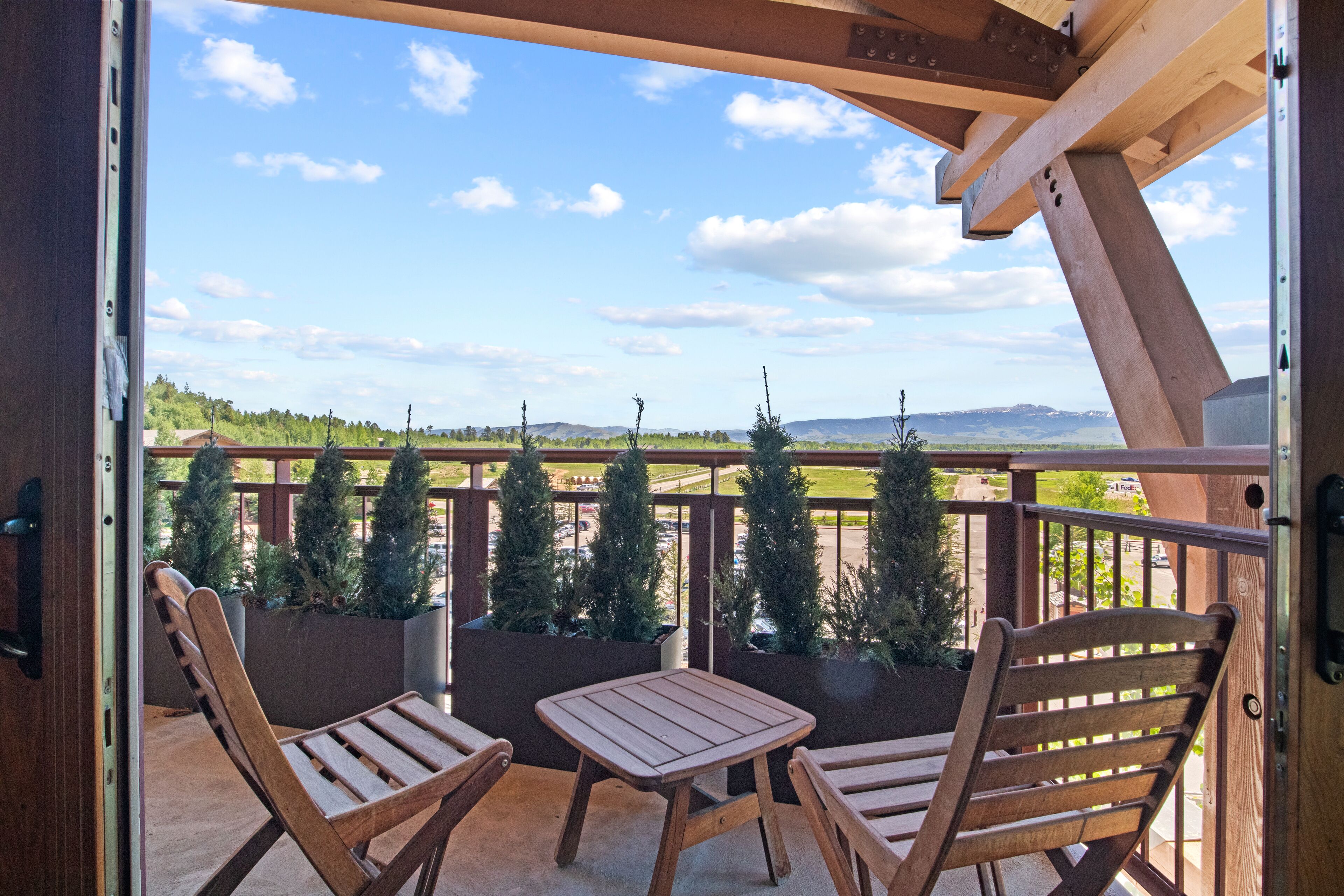 Een balkon met houten stoelen en een tafel, met uitzicht op een aangelegd gebied met bomen en heuvels in de verte.