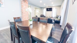 Een eethoek met een houten tafel en stoelen, een wand van baksteen en zicht op een woonkamer met een bank en televisie.