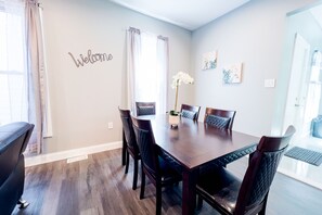 A dining area with a wooden table and chairs, a 'Welcome' sign on the wall, and artwork. - The Haines Villa (Niagara Falls)