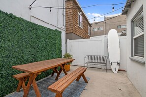 Una mesa de picnic de madera con bancos, una tabla de surf blanca apoyada contra una pared y un seto.
