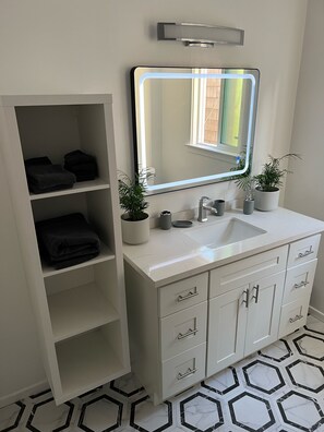 Un baño con un tocador blanco, un espejo grande, dos plantas en macetas y un suelo con estampado hexagonal.