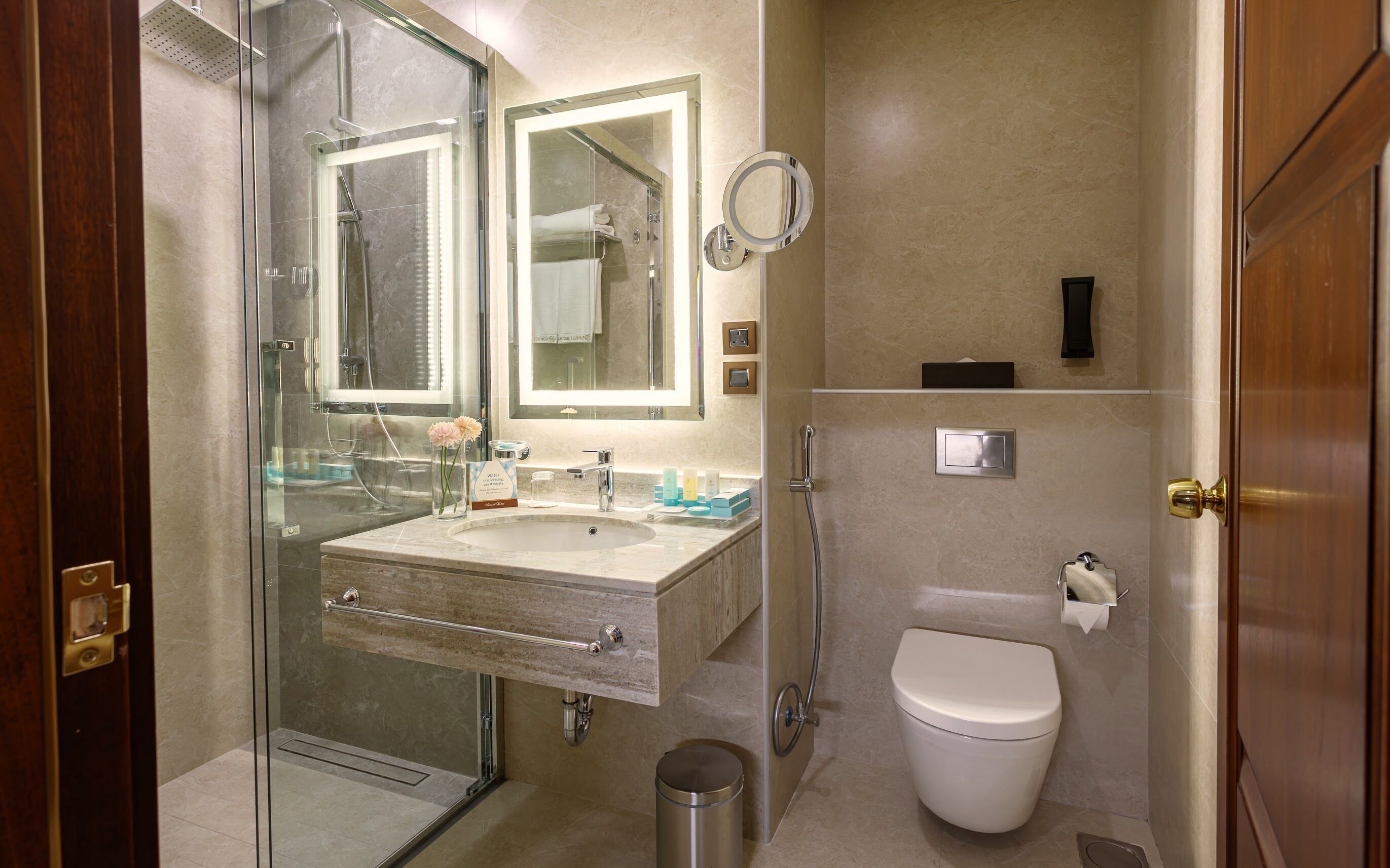 Un baño moderno con ducha envuelta en vidrio, lavamanos con tocador, inodoro y espejo con luz de aro.