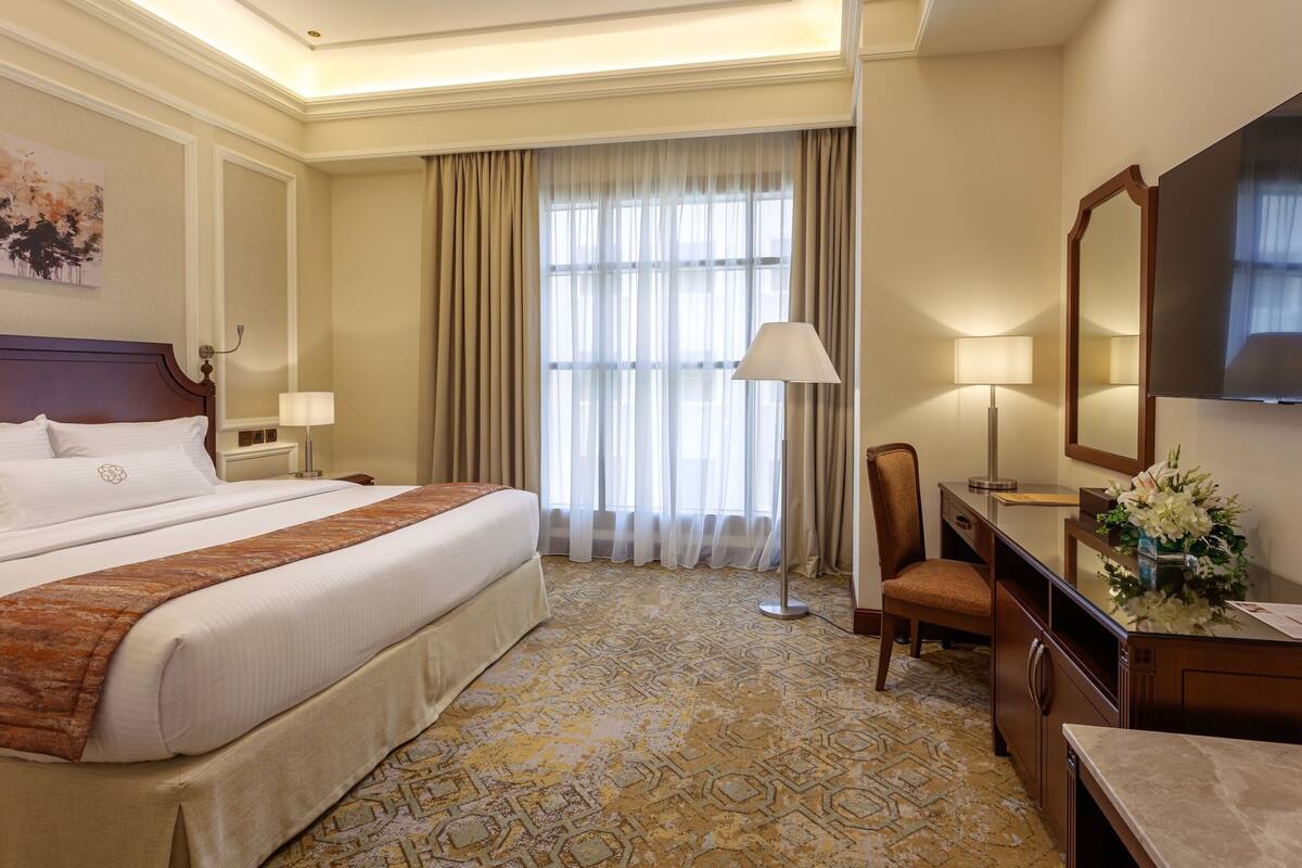 Habitación de hotel con una cama grande, un escritorio con silla, una lámpara de escritorio, un espejo y una ventana con cortinas.
