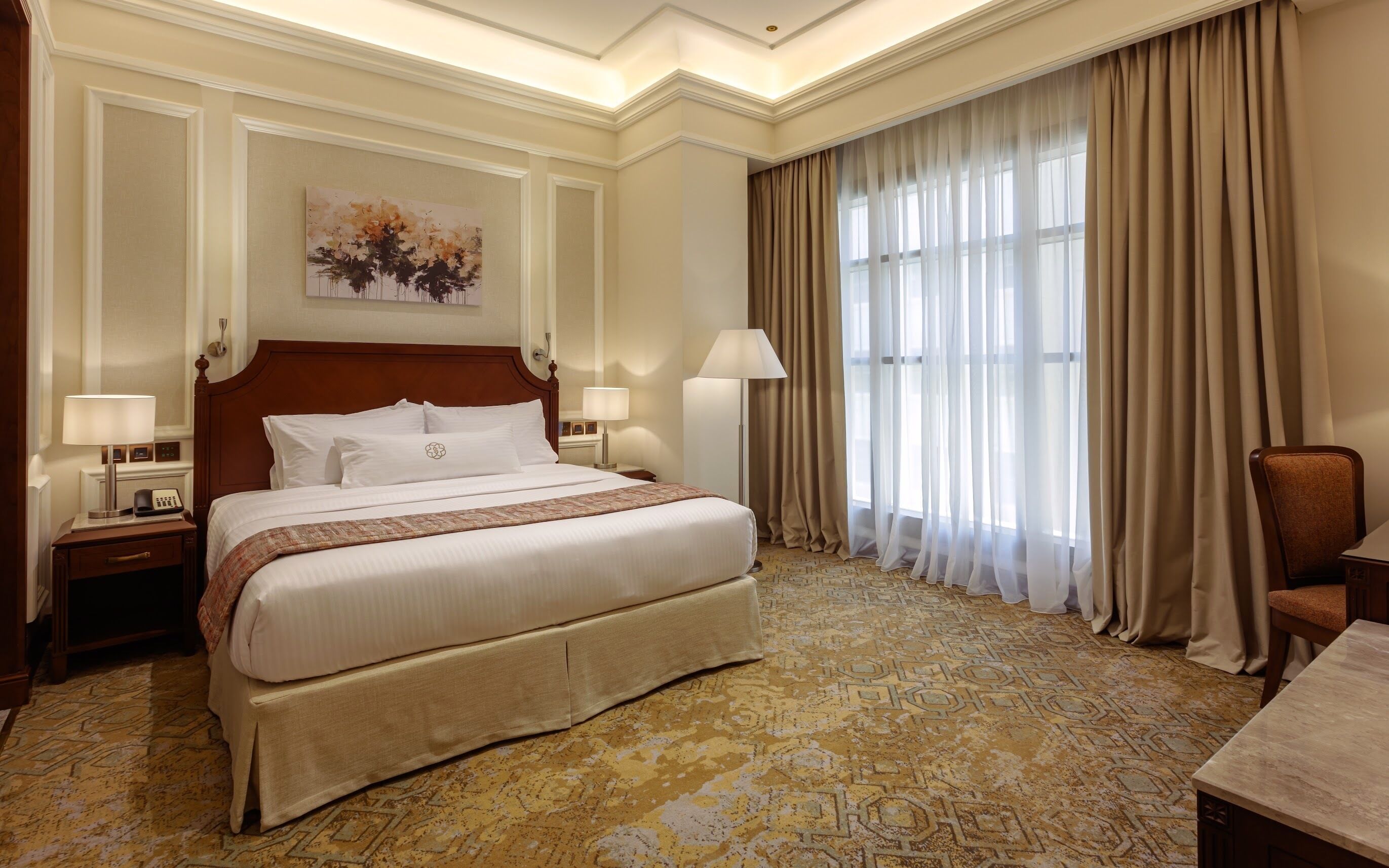 A hotel room with a large bed, two bedside tables with lamps, a large window with sheer curtains, and a carpeted floor.