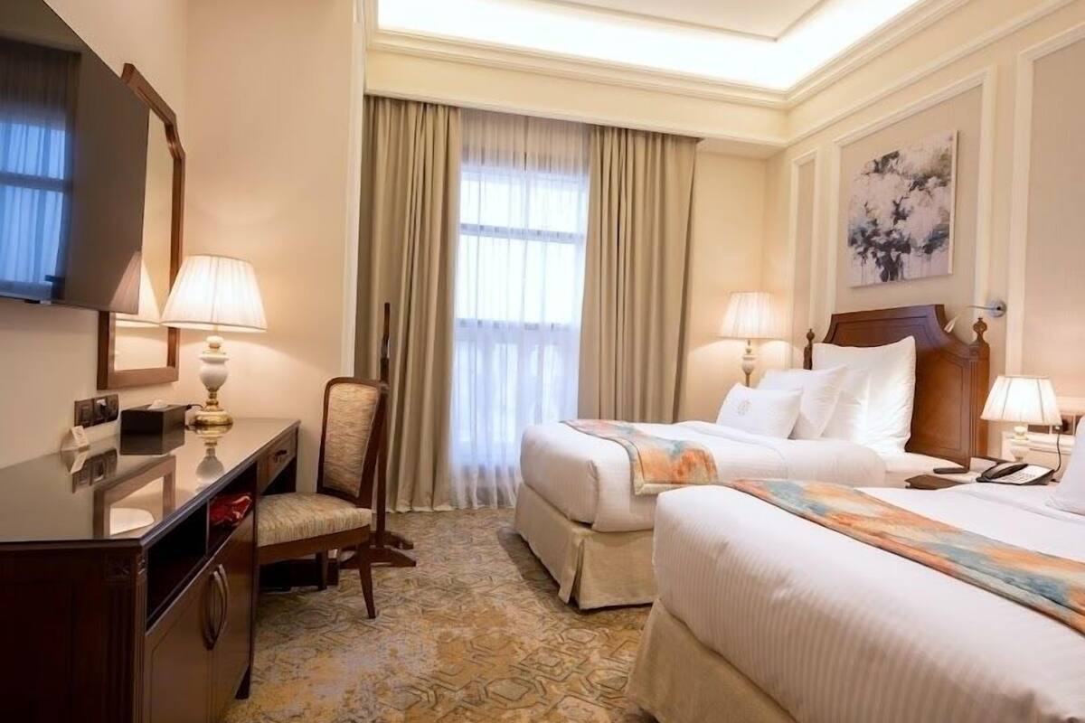 Habitación de hotel con dos camas, un escritorio, una silla, un televisor y un ventanal con cortinas.