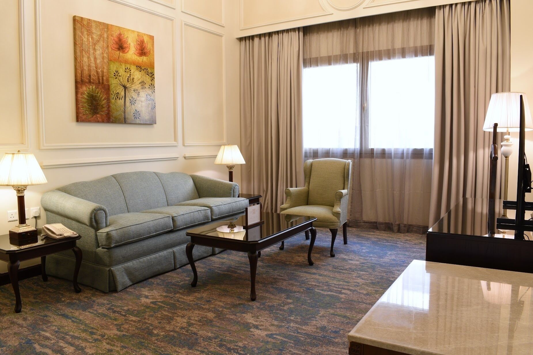 Habitación de hotel con sofá verde, un sillón a juego, una mesita y un escritorio con lámpara. Se aprecia un ventanal con cortinas, un cuadro en la pared y suelo alfombrado.