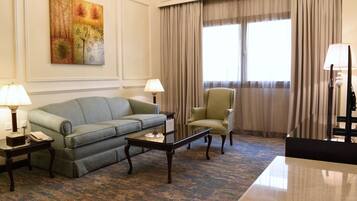Habitación de hotel con sofá verde, un sillón a juego, una mesita y un escritorio con lámpara. Se aprecia un ventanal con cortinas, un cuadro en la pared y suelo alfombrado.