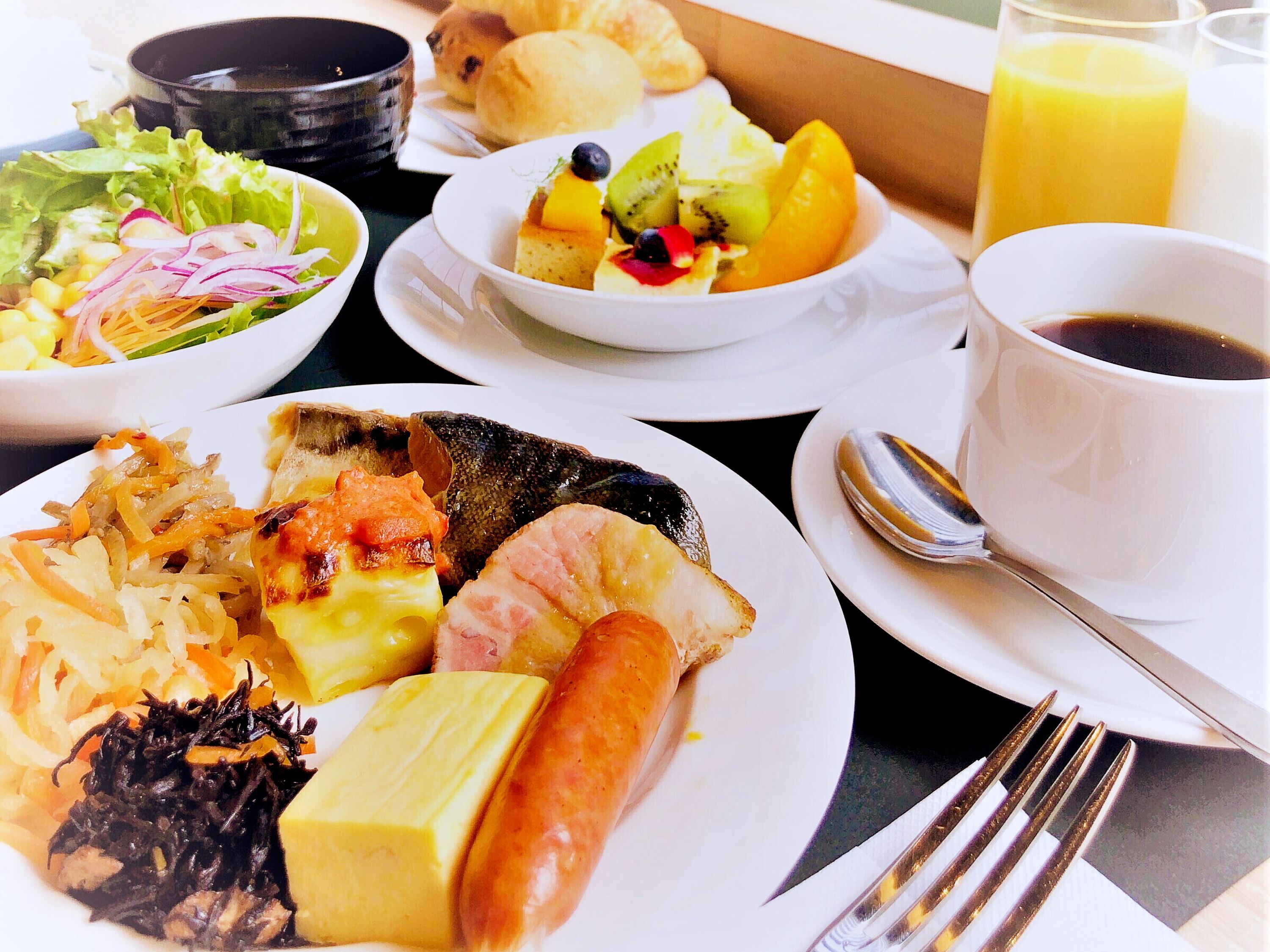 A table with various dishes including a salad, a fruit bowl, a plate with sausage and vegetables, and a cup of coffee.