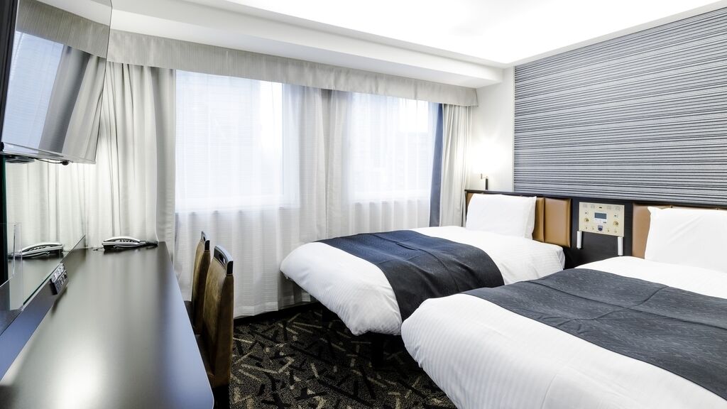 A hotel room with two beds, a desk, a TV, and a window with curtains.
