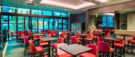 A modern restaurant with red upholstered chairs and tables, large windows offering a view of the street, and a bar area with a counter and stools.