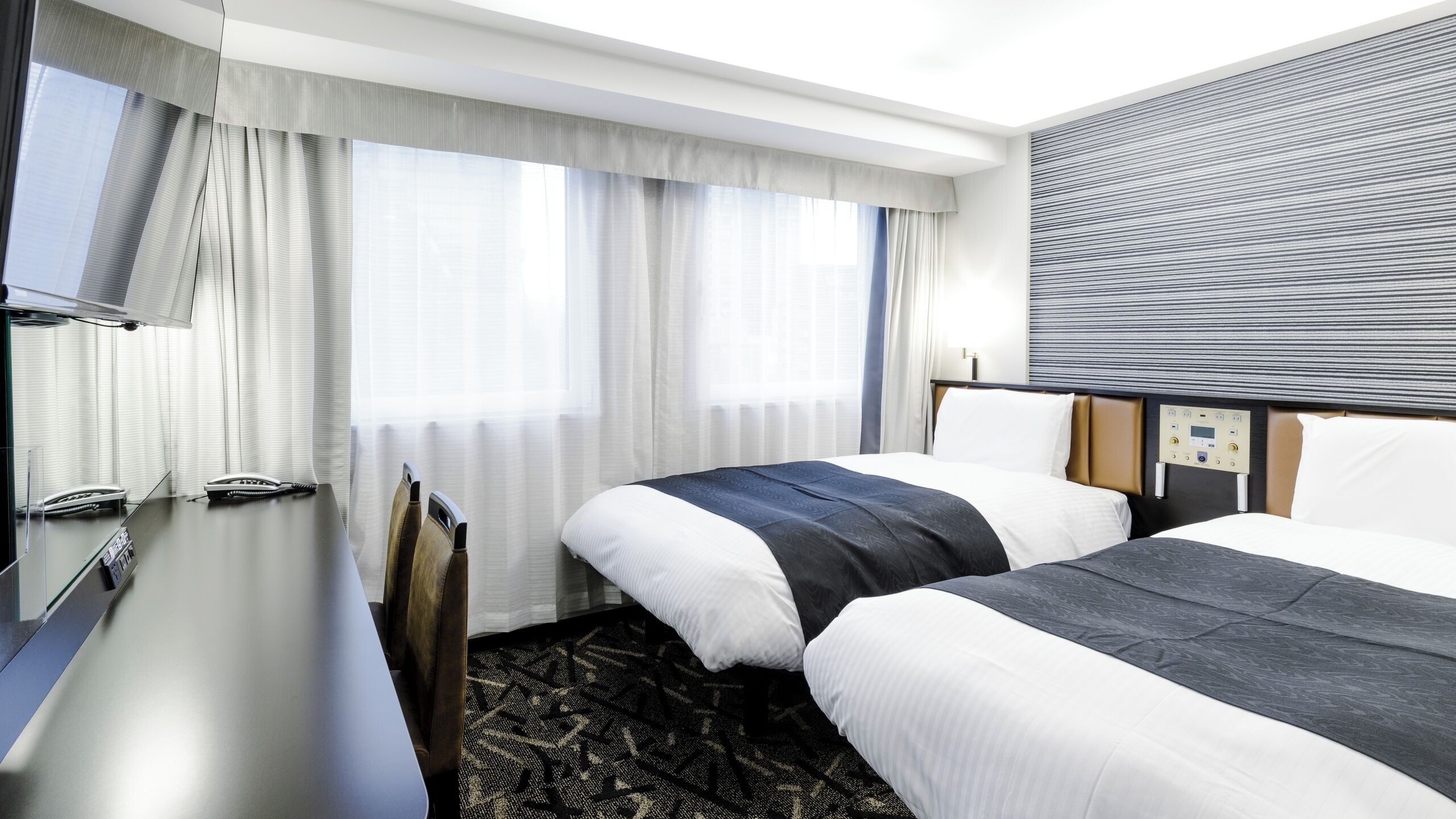 A hotel room with two beds, a desk, a television, and a control panel on the wall.