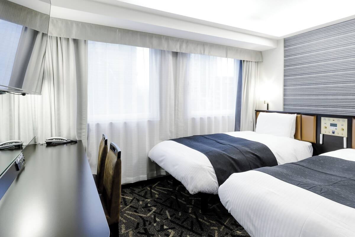 A hotel room with two beds, a desk, a television, and a control panel on the wall.