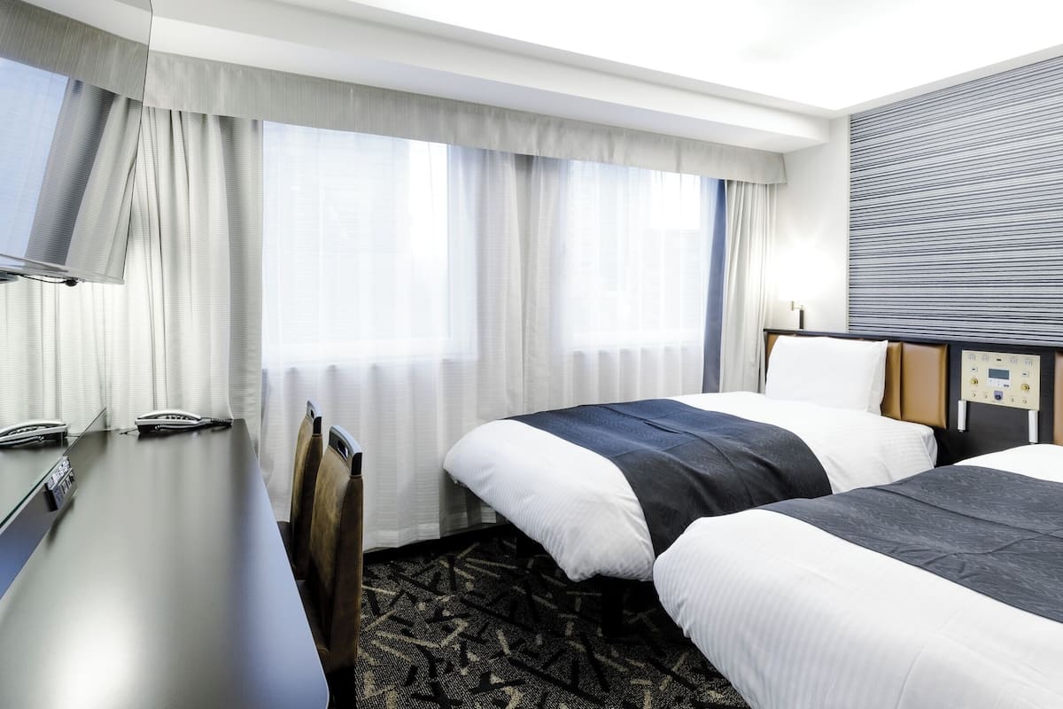 A hotel room with two beds, a desk, a television, and a control panel on the wall.