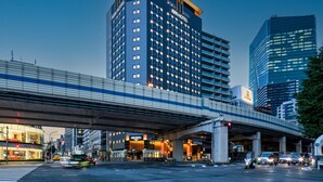 A modern hotel with a prominent 'APA HOTEL' sign, located under an overpass in an urban setting with visible traffic and other buildings. - APA Hotel & Resort Roppongi Ekihigashi (Tokyo)