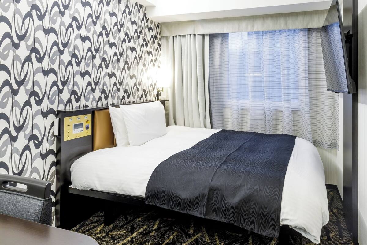 A hotel room with a bed, a control panel, curtains, and a patterned wall.