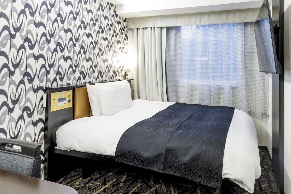 A hotel room with a bed, a control panel, curtains, and a patterned wall.