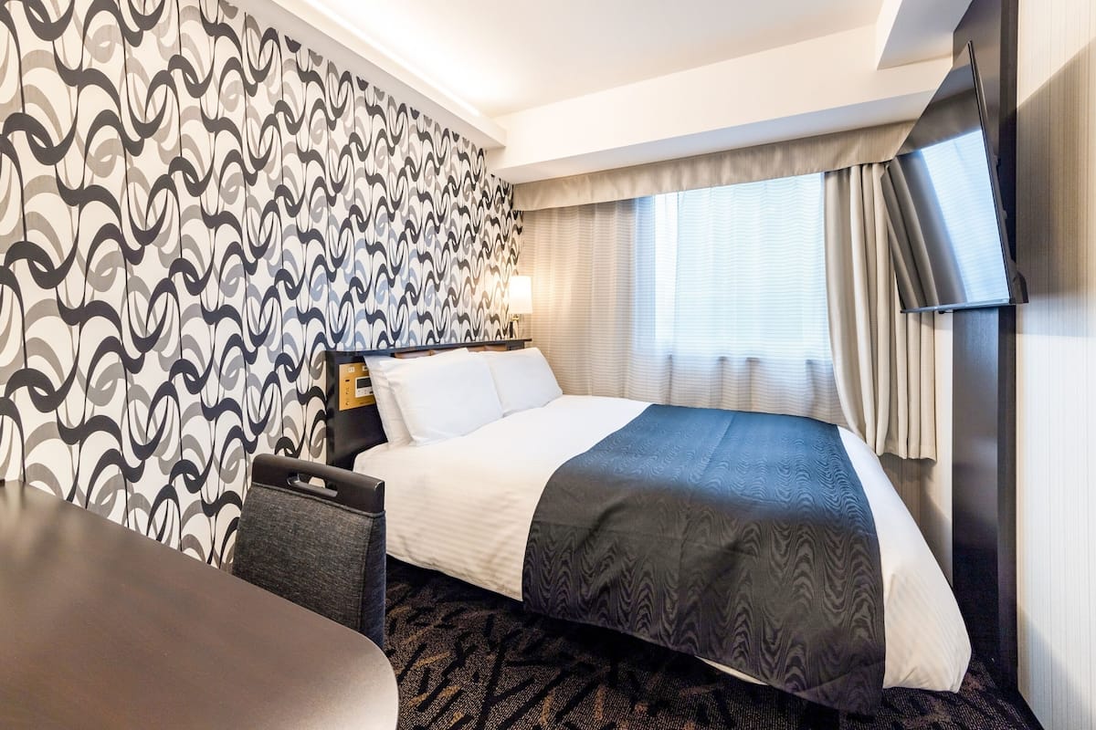 A hotel room with a bed, a desk, a chair, a TV, and a patterned wall.