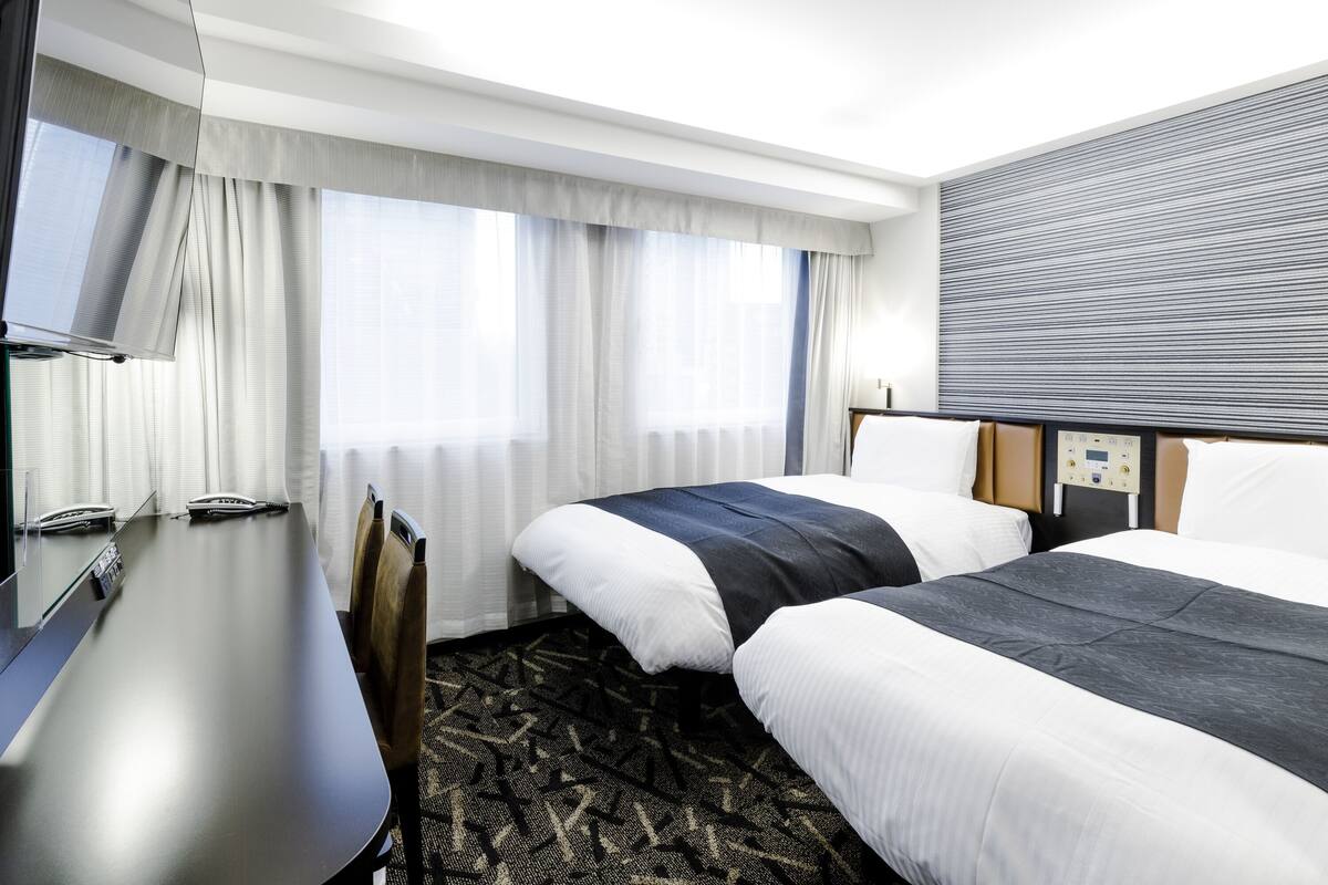 A hotel room with two beds, a desk, a chair, a television, and a control panel on the wall.