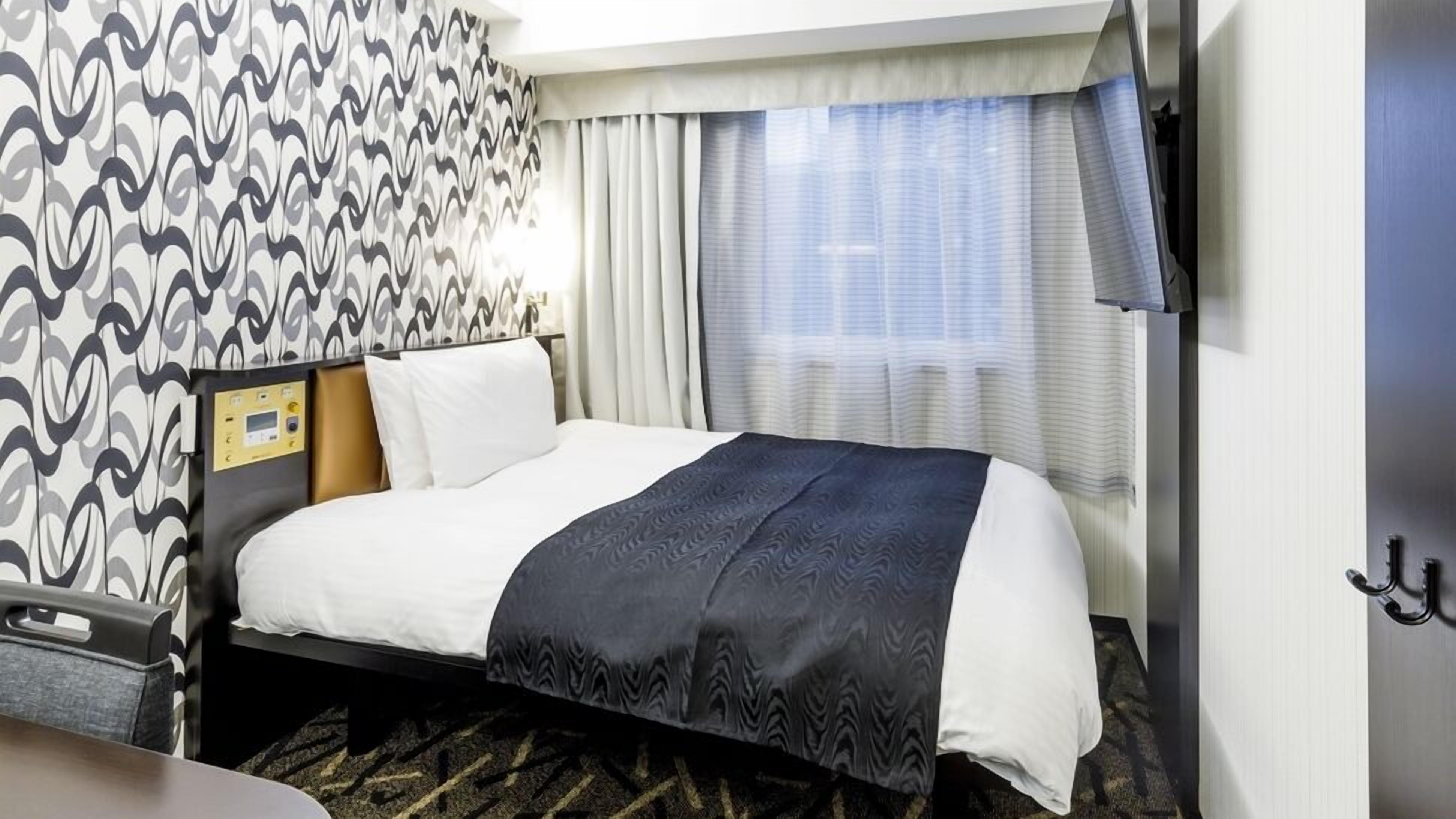 A hotel room with a bed, a control panel, a window with curtains, and a patterned wall.
