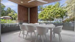 Uma área de refeições moderna ao ar livre, com mesa e cadeiras brancas, painéis de madeira e vista para vegetação exuberante e um corpo d'água.