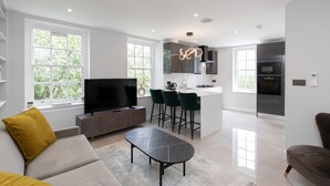 Uma sala de estar moderna com uma TV de tela plana, um sofá, uma mesa de centro e uma cozinha com banquetas.
