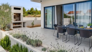 A modern outdoor patio with a dining table and chairs, a built-in grill, and a landscaped garden with various plants. - Vacation home Inizio by Interhome (Brodarica)