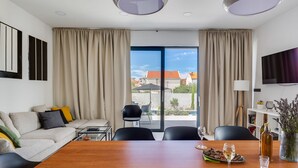 A modern hotel room with a large wooden dining table, a sofa with colorful cushions, a TV mounted on the wall, and a balcony with a view of the city. - Vacation home Inizio by Interhome (Brodarica)