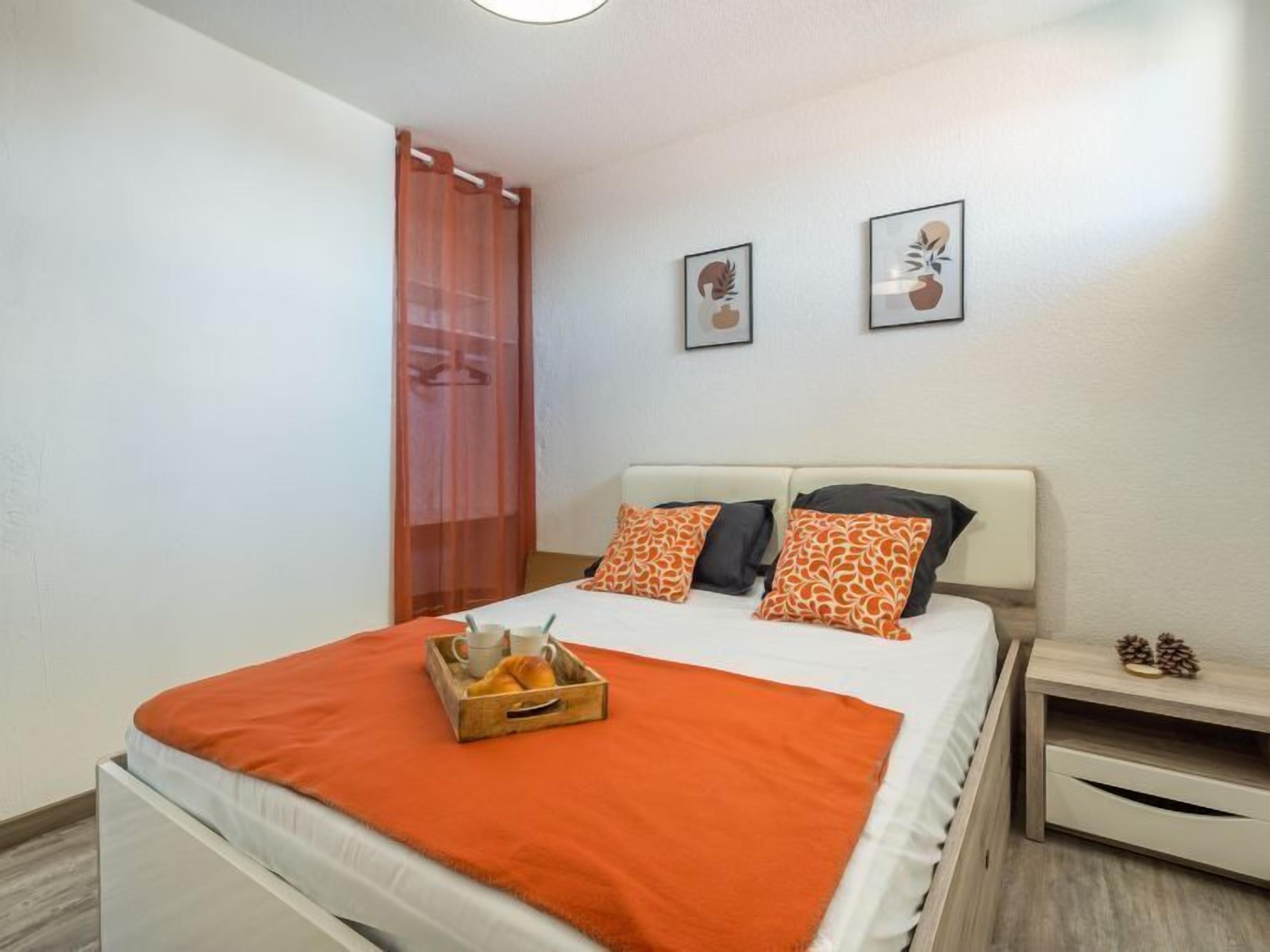 Un dormitorio con cama, mesita de noche, mesita auxiliar de madera y ventana con cortina ligera.