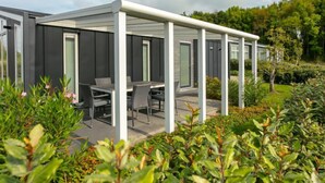 Un espace extérieur moderne, aménagé en patio, avec un coin salon abrité, entouré d’une végétation luxuriante et surplombé d’un ciel dégagé.