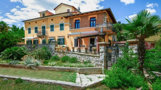 Una casa de dos pisos con un balcón, rodeada de vegetación y un muro de piedra.