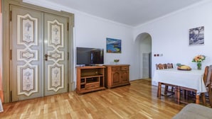 Una habitación con piso de madera, un televisor sobre un mueble de madera, una mesa de comedor con sillas y un guardarropa verde con paneles decorativos.