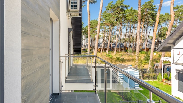 A modern building with a glass balcony, a metal railing, and a view of tall trees and other buildings in the background.