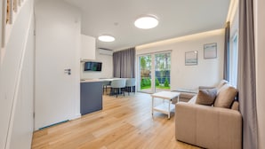 A modern living room with a sofa, a small table, and a TV. There is a kitchen area with a dining table and chairs. The room has wooden flooring and large windows with curtains.